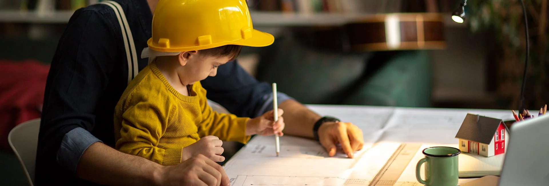 Ein kleiner Junge mit gelbem Helm sitzt bei seinem Vater auf dem Schoß an einem Schreibtisch. Der Vater zeigt dem Jungen einen Bauplan.