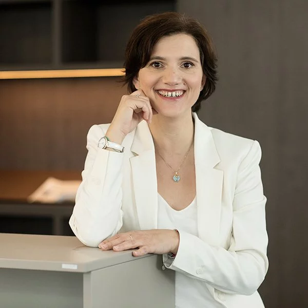 nyoo-Architektin Anna Maria Conoci trägt einen weißen Blazer und steht lächelnd, angelehnt an einem kleinen Schrank in einem Büro.