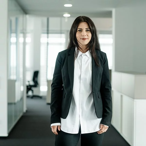 Jessica Machado aus dem Team "Bauzeichnung" bei nyoo steht in einem schwarzen Anzug in einem Büro-Flur.