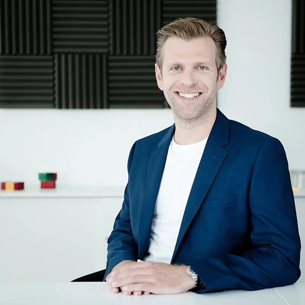 Michael Hannen aus dem Team "Development" bei nyoo trägt einen dunkelblauen Anzug und sitzt lächelnd an einem Bürotisch.