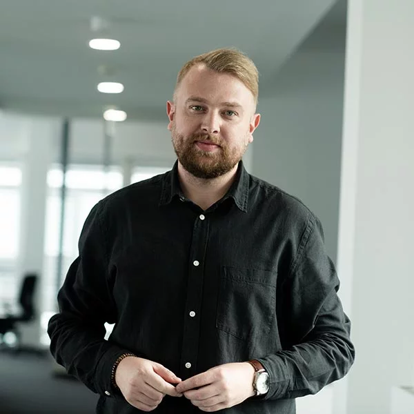 Planer für Architektur Alexander Weissheim steht in einem schwarzen Hemd in einem Büro und lehnt sich mit einem Ellenbogen an einen kleinen Schrank und lächelt.