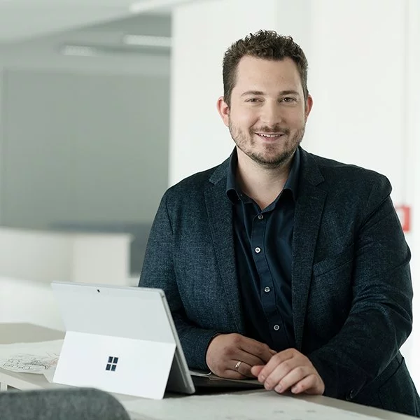 Simon Bruckschen aus dem Team "Planungskoordination" bei nyoo trägt einen dunkelgrauen Anzug und steht lächelnd an einem Bürotisch mit einem Tablet vor ihm.