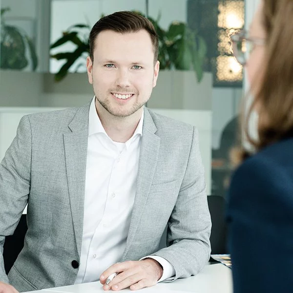 nyoo-Projektentwickler Johannes Dangelmayer sitzt in einem grauen Anzug an einem Bürotisch und lächelt.